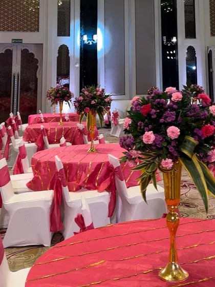Bright and cheerful table settings for a Mehendi function. The tables are dressed in vibrant pink linens with gold details, topped with tall floral centerpieces in matching hues.