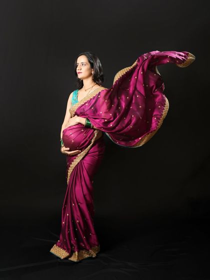 A dramatic and elegant maternity photo featuring a flowing magenta saree against a dark backdrop. This style of shot emphasizes the beautiful silhouette and the rich colors of the traditional fabric.