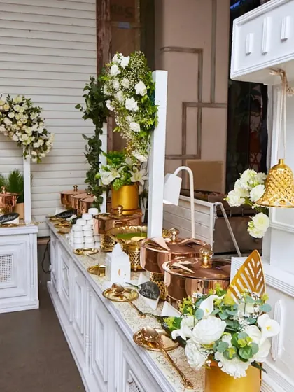 An elegant white and gold buffet setup. The copper serving pots, golden leaf decor, and a hanging brass bell add a touch of traditional luxury.