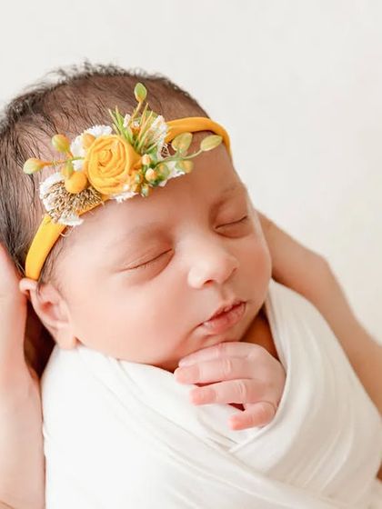 A close-up shot of a newborn's face, held gently by their parent's hands, focusing on their delicate features and yellow floral headband.