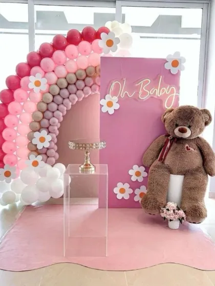 A fun and sweet baby shower backdrop featuring a rainbow balloon arch made of pink and neutral tones. The design is accented with cute daisy cutouts, an 'Oh Baby' neon sign, and a friendly teddy bear.
