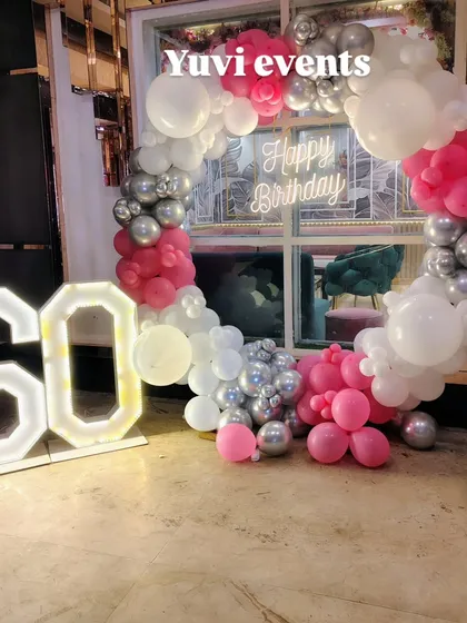 A fun and vibrant setup for a 60th birthday, using a pink, silver, and white balloon ring with a neon sign and marquee numbers.