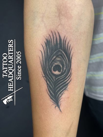 A black and grey peacock feather tattoo on the forearm, with detailed shading to create a realistic and soft texture.