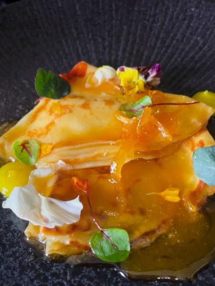 A delicate crêpe suzette, folded and served with a vibrant orange sauce and edible flowers. A timeless French dessert.
