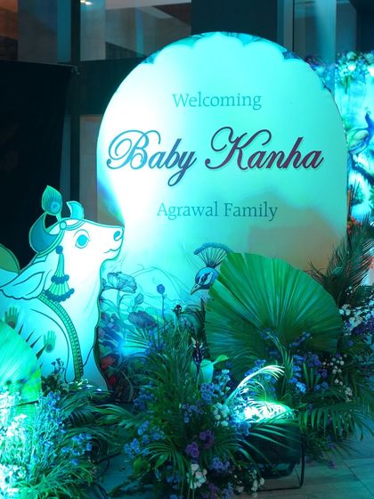 The welcome sign for the 'Baby Kanha' baby shower. The sign is set against a backdrop of tropical leaves and blue florals, with a beautiful cutout of a sacred cow, introducing the divine theme.