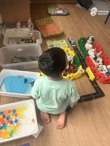 A city is being built, one sentence at a time. This setup invites a child to explore different sensory bins, from water beads to sand, creating a rich context for language to flourish naturally through play.
