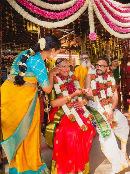 The sister of the bride assisting during a Tamil wedding ceremony. Her bright yellow and blue Kanjeevaram saree is a classic choice, looking beautiful against the bride's red silk.