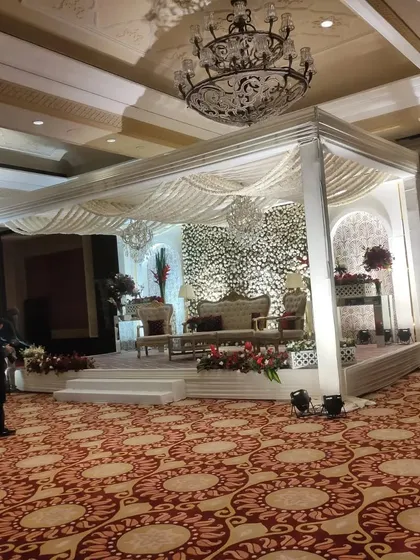 An elegant and grand indoor wedding stage setup within a banquet hall. The design features a pristine white mandap structure, a dense floral backdrop of white flowers, and luxurious seating, all under a classic crystal chandelier.