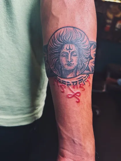 A front view of the 'Har Har Mahadev' armband tattoo. The red calligraphy stands out against the black and grey portrait, making the chant a central focus of the design.