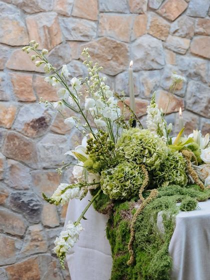 A detailed shot of the floral arrangement on the Earth Table. Tall white delphiniums and green hydrangeas create height and texture, with moss spilling over the side of the table.