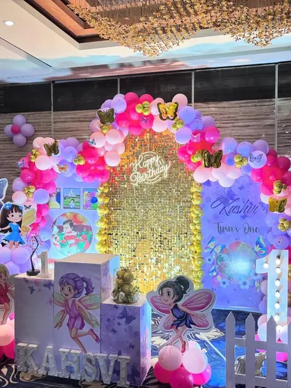 A magical fairy-themed first birthday party. The centerpiece is a gold sequin shimmer wall, framed by a pink, purple, and lilac balloon arch, and accented with fairy character cutouts and light-up marquee letters.