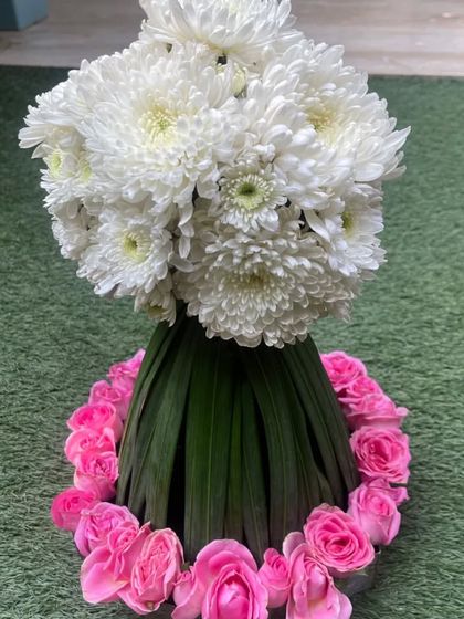 A beautiful floral creation featuring a base of folded green leaves, a ring of pink roses, and a crown of white chrysanthemums. This is a wonderful example of a structured yet soft centerpiece.