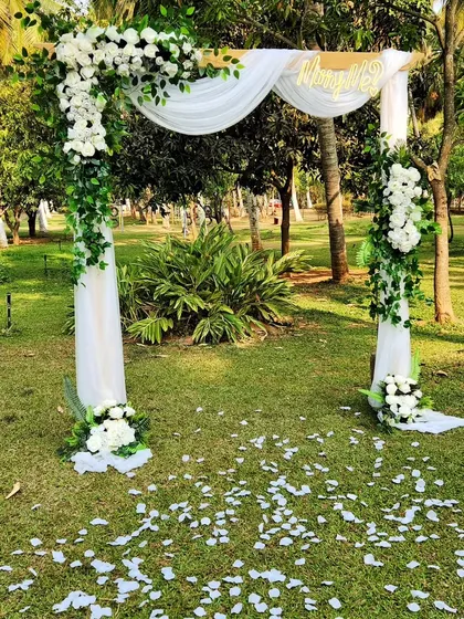 A simple and elegant daytime proposal arch set in a lush green park. The wooden structure is draped with white fabric and adorned with white roses and greenery, with petals scattered on the grass below.