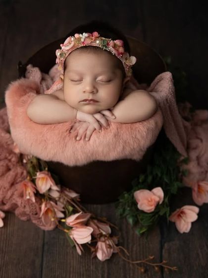 This baby girl looks like a sleeping fairy, nestled in a bucket with soft pink fabrics and surrounded by delicate flowers on a dark wood floor.