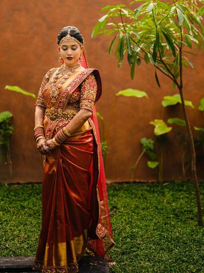 A full-length shot of the bride in a garden setting. The vibrant red of her saree stands out beautifully against the green foliage.