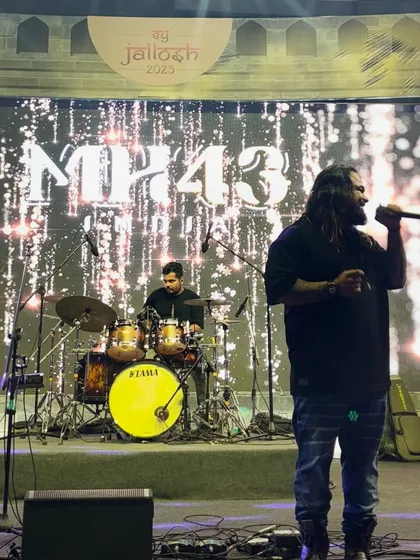 A wide shot of the stage at a corporate event, showing our frontman and drummer against the "MH43" LED screen.