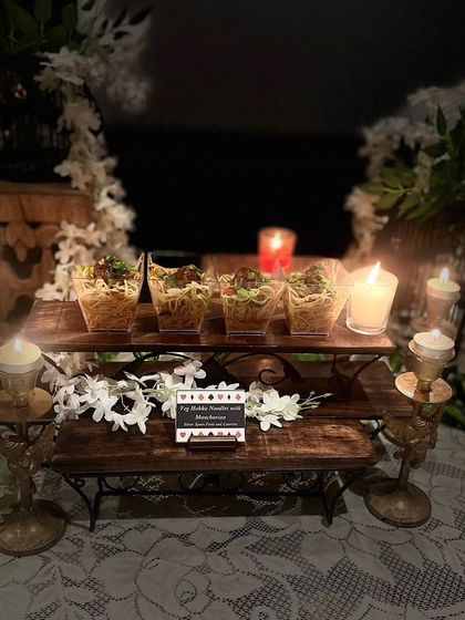The Veg Hakka Noodles display in a darker, more intimate lighting setting. The candles create a warm glow, highlighting the food and the delicate floral decorations.