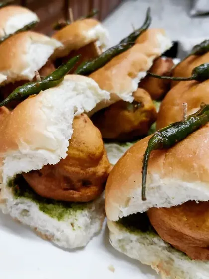 A close-up of our mini Vada Pavs. Each soft bun is layered with spicy green chutney and holds a perfectly fried potato vada, garnished with a fried green chili for that authentic touch.