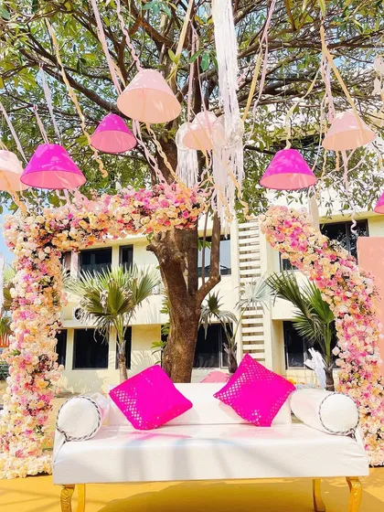 This special seating area for a Haldi ceremony is set under a tree decorated with hanging lampshades. The floral arch and bright pink cushions create a cheerful and picturesque spot for the couple.