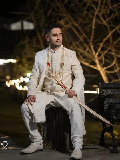 A full-length portrait of the groom seated outdoors at night. The lighting creates a dramatic, cinematic feel, highlighting his sharp attire.