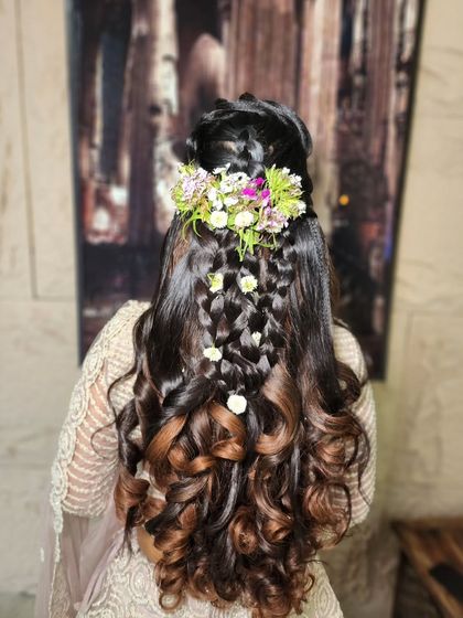 An elegant and modern hairstyle with intricate braids and soft curls, adorned with fresh flowers. This is a perfect look for an engagement or sangeet ceremony.