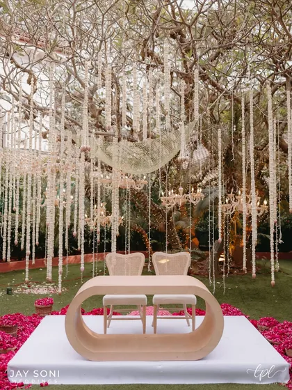 A modern and romantic mandap setup under a canopy of hanging floral strings and chandeliers. The unique, curved wooden table and minimalist chairs create a chic and intimate space for the wedding ceremony.
