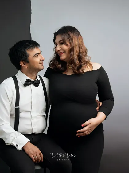 An elegant and formal studio maternity portrait of a couple. The man, in a bow tie and suspenders, looks lovingly at his pregnant partner, who is dressed in a classic black gown.