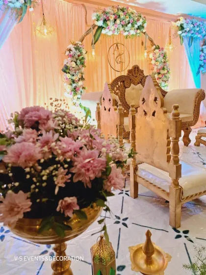 A detailed shot of the seating area for an engagement ceremony. The setup includes traditional wooden chairs for the rituals, placed in front of the main stage, with brass flower vases and lanterns adding to the decor.