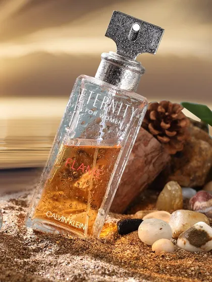 A creatively styled product shot of a Calvin Klein perfume bottle on a bed of sand and pebbles. This composition tells a story of summer, beaches, and eternity, perfectly matching the product's name.