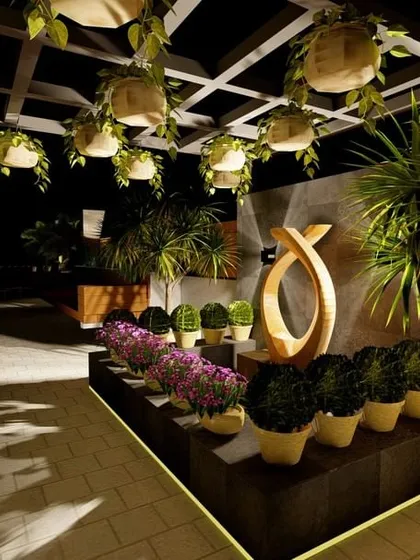 A beautifully arranged planter bed on a terrace, featuring a mix of flowering plants and sculptural greenery under a pergola with hanging plants. The design includes a unique wooden artifact, adding an artistic touch.