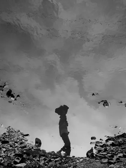 The reflection of a worker carrying bricks is captured in a puddle, an artistic black and white composition that offers a different perspective on their daily toil.
