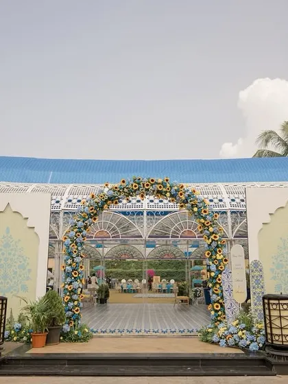The grand entrance to the glasshouse for the Jodhpur-themed Haldi. A floral arch of blue and yellow flowers welcomes guests into a space filled with custom prints and vibrant colors.