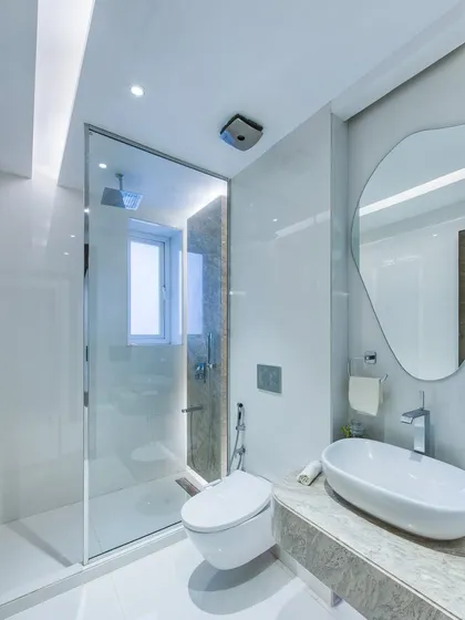 A bright and clean bathroom design with white marble walls and a frameless glass shower enclosure. The organic-shaped mirror adds a soft, contemporary touch to the minimalist space.