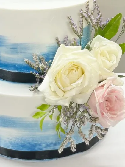 A two-tier cake with a beautiful blue watercolor effect, accented with fresh white and pink roses. The black ribbon adds a touch of modern contrast.
