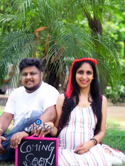 A relaxed portrait of the couple sitting in a park for their maternity announcement photoshoot, holding a "Coming Soon" sign.