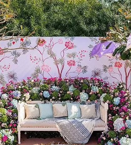 A beautiful photo backdrop for a Mehendi or garden party. It features a large canvas with a delicate floral print, with a vintage white bench and a lush border of real flowers in front.