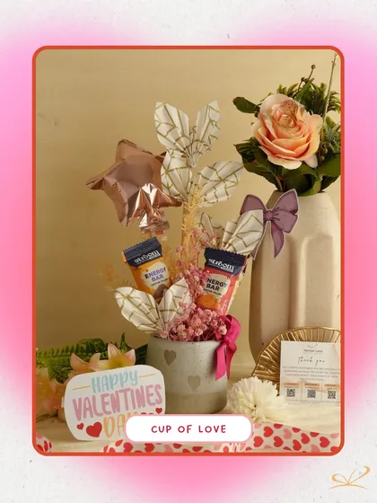 The 'Cup of Love' hamper is a compact and cute arrangement in a mug. It includes energy bars, a small balloon, and decorative elements, making it a perfect small token of affection.