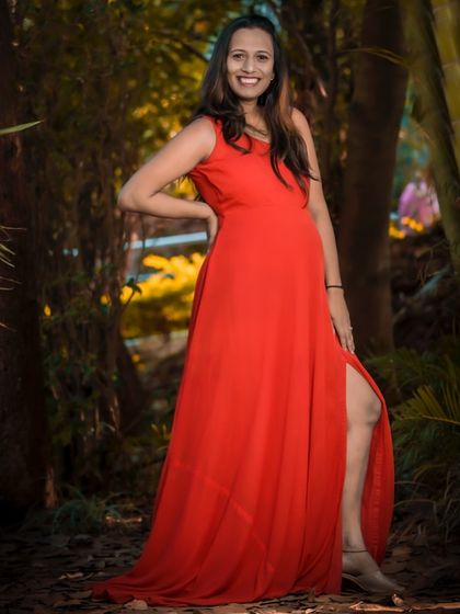 A confident and glamorous solo portrait in a striking red gown. The pose highlights both the baby bump and the elegance of the dress, set against a beautiful, warmly lit natural background.