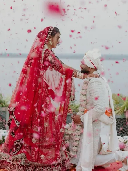 A romantic varmala moment with a shower of pink rose petals. The bride is in a stunning red lehenga, and the decor is set against a serene water body, making the entire scene feel like a beautiful painting.