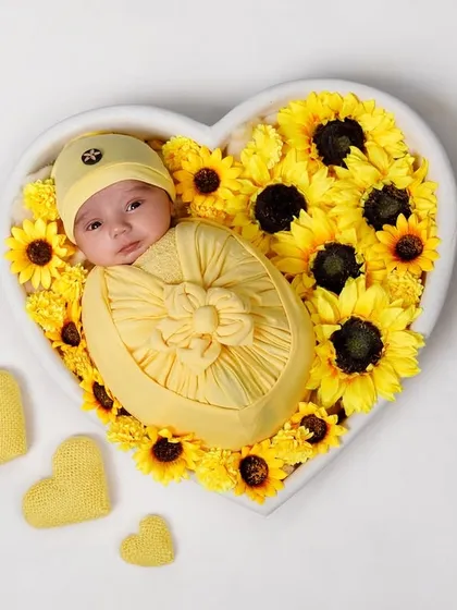 A newborn boy lies awake in a heart-shaped bowl filled with bright yellow sunflowers.