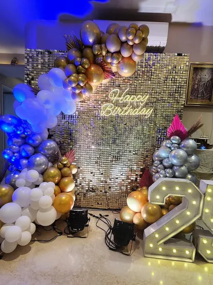 A chic and stylish 25th birthday party decoration. The centerpiece is a gold shimmer wall with a neon sign, framed by a modern balloon arch in gold, silver, and white, and accented with large light-up marquee numbers.