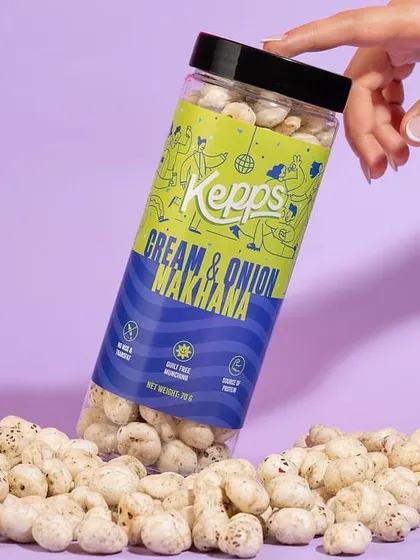 A tilted shot of a Cream & Onion Makhana jar, with the snacks spilling out onto a purple surface. The hand interacting with the jar adds a human element, making the product feel accessible and ready to be enjoyed.
