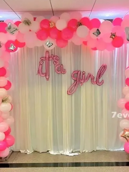 A lovely 'It's a Girl!' announcement or baby shower backdrop. A pink and white balloon arch frames a simple curtain, with foil letters sharing the happy news.