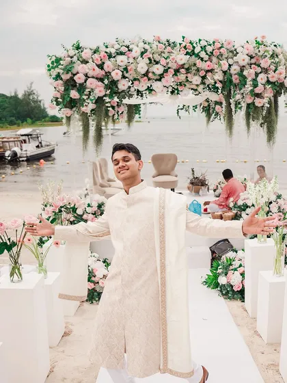 The groom, full of joy and excitement, stands at the floral mandap on a beach in Thailand, ready to welcome his bride. This candid shot perfectly captures the happy and relaxed vibe of a destination wedding.