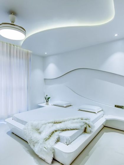 A futuristic and minimalist bedroom designed entirely in white. The custom-built bed platform flows seamlessly into the wavy, sculptural wall and ceiling, creating a fluid and serene space.