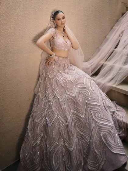 A bride in a blush pink lehenga, wrapped in sparkle and grace. The look is completed with a delicate sheer veil that whispers elegance with every step.
