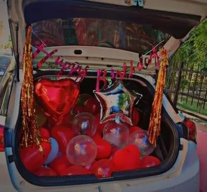 A romantic car boot surprise. The trunk is filled with red heart-shaped balloons and a 'Happy Birthday' banner, perfect for a celebration on the move.