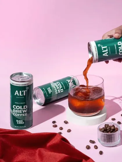 This action shot of cold brew being poured into a glass is both dynamic and refreshing. The vibrant pink background contrasts beautifully with the dark coffee and green can, creating a modern and eye-catching image.