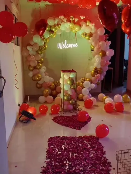 A stylish welcome setup featuring a circular balloon arch in red, white, and gold. A "Welcome" neon sign and a rose petal pathway complete the look.