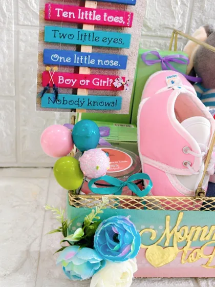 A close-up of the details in a gender-reveal hamper, showing the decorative sign and tiny pink booties. Every element is chosen to fit the "Boy or Girl? Nobody knows!" theme perfectly.